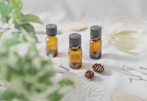 3本の茶色いエッセンシャルオイルボトルが白いテーブルの上に並んでおり、ボトルの周りには植物や松ぼっくり、小枝が飾られている。